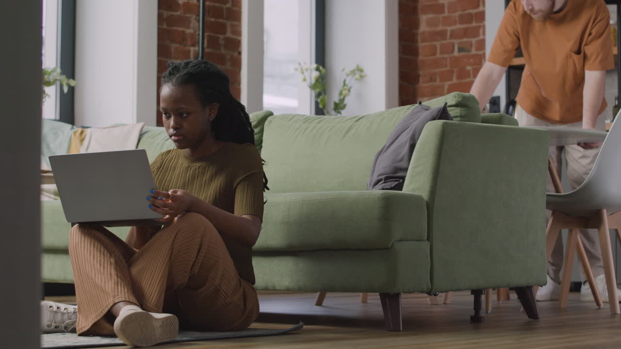 Girl Working On Laptop Computer Sitting On Floor While Her Male Roommate Cleaning The Kitchen Table