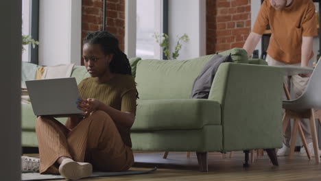 girl working on laptop computer sitting on floor while her male roommate cleaning the kitchen table