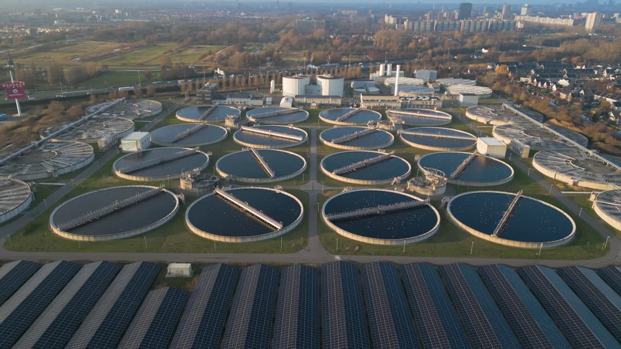 Drone approach shot flying toward large operational wastewater clarifier basins at the Harnaschpolder treatment plant in the Netherlands with sunlight reflections across water surfaces