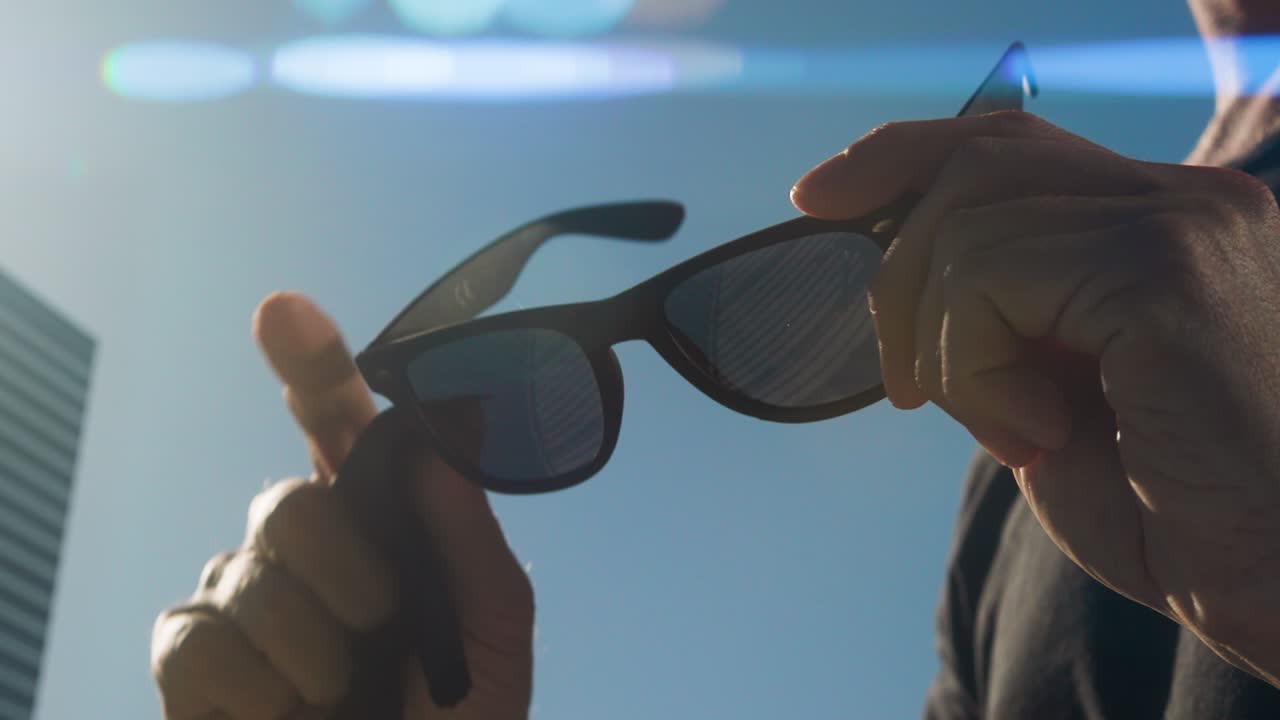 검은 선글라스의 오른쪽 렌즈를 는 손, 검은 티셔츠를 입은 남자의 몸통, 파란 하늘과 배경의 건물, 은 날씨, 빛의 아나모프스 플레어