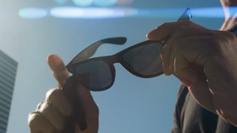 Close-up-of-hands-polishing-the-right-lens-of-black-sunglasses,-torso-of-man-in-black-t-shirt,-blue-sky-and-building-in-the-background,-sunny-weather,-anamorphic-flares-from-sunlight