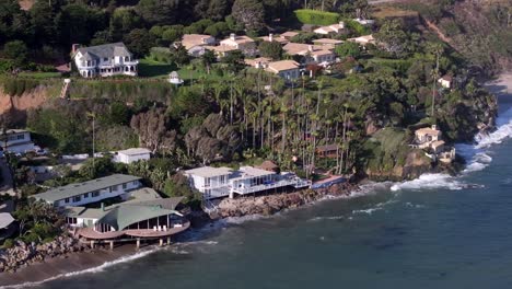 luxury beach front multi million dollar homes over malibu pacific ocean coastline, aerial rising on picturesque day while waves crash along the shore