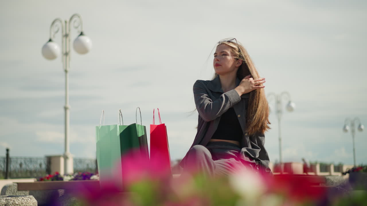 mujer joven sentada en un banco al aire libre con bolsas de compras a su lado, ajustando su cabello a medida que sopla el viento, tiene gafas de sol descansando en su cabeza y se sienta en una postura relajada en un día soleado