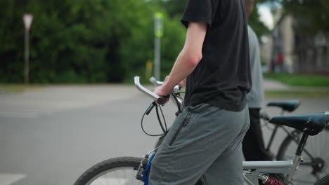 two brothers walking side by side, each pushing their bicycles across a pedestrian crossing, the background features a signpost and blurred greenery and a residential building in the distance