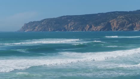 Azure-waves-white-with-foam-in-the-ocean-with-a-rocky-cape-in-the-background,-Portugal