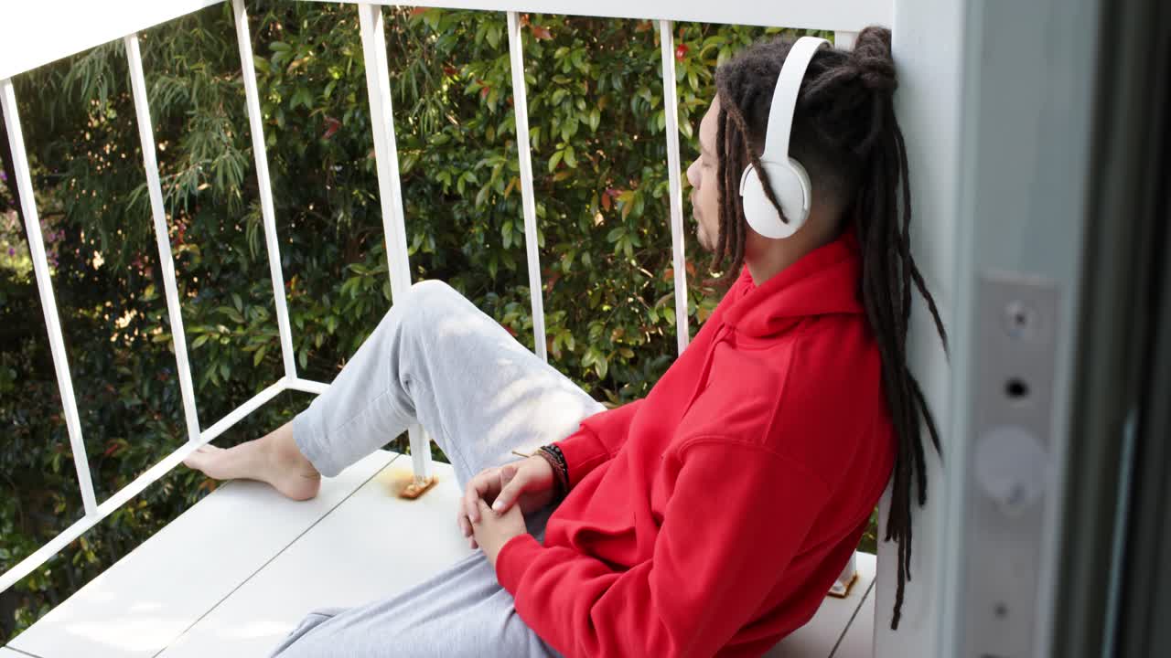 Ripresa panoramica con una fotocamera di un adulto con cappuccio rosso che mette le cuffie sul balcone, mentre guarda il fogliame verde ascoltando