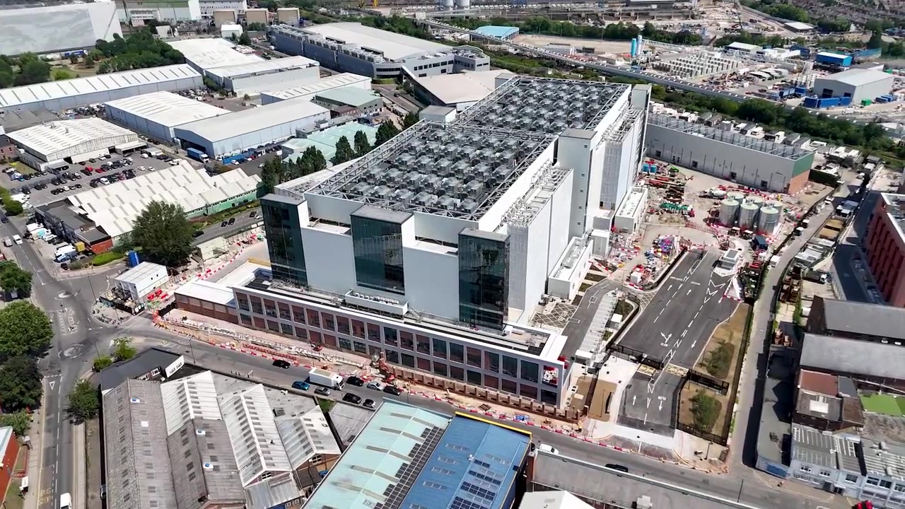 Edificio de centro de datos con planta de refrigeración en la azotea se eleva sobre un polígono industrial del oeste de Londres con almacenes y patios, con vallas de obra a lo largo de la carretera.