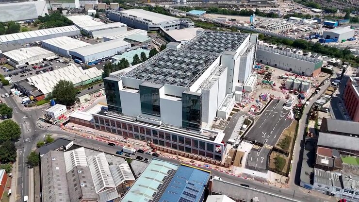 Data centre building with rooftop cooling plant rises above a west London industrial estate of warehouses and yards, with construction hoardings along the road