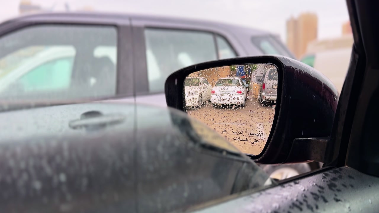 Close-up of a rain-covered car side mirror reflecting parked vehicles on a wet urban street, capturing moody weather, roadside traffic, and everyday city driving in soft overcast light.