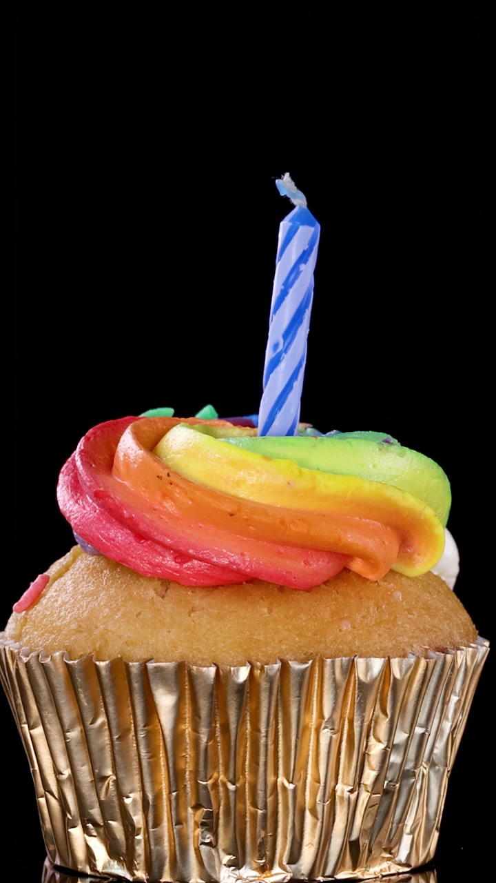 Una mano inserisce con cura una candelina di compleanno a strisce blu in un cupcake con glassa arcobaleno e pirottino dorato, su uno sfondo nero con illuminazione brillante e uniforme