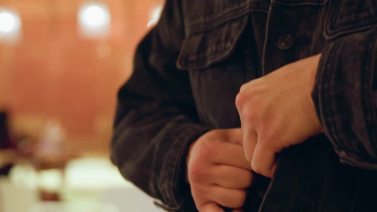 Close up of man buttoning his dark denim jacket with both hands visible focusing on action and texture details. background is softly blurred indoor setting with warm lighting suggesting