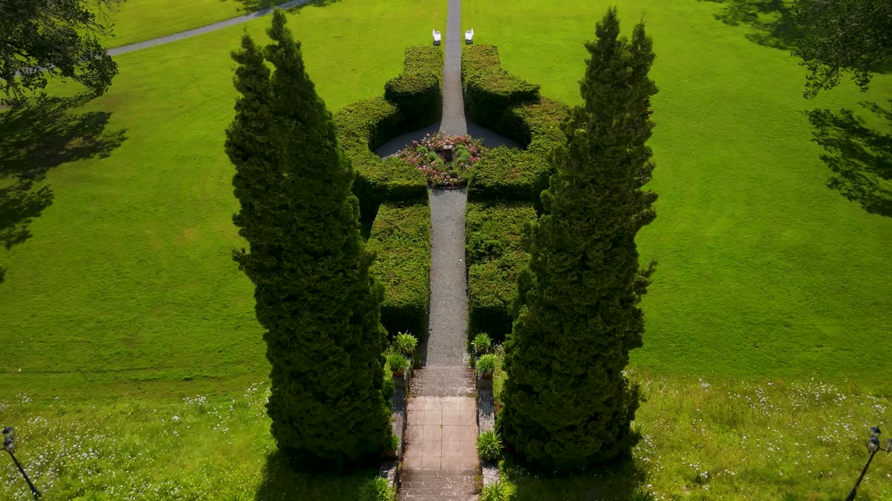 Eine Drohnenkamera schwenkt nach oben und enthüllt einen symmetrischen Gartenweg mit gestutzten grünen Büschen und perfekt gepflegten Rasenflächen auf dem historischen Gutshof Baldersnäs Herrgård in Dalsland, Schweden