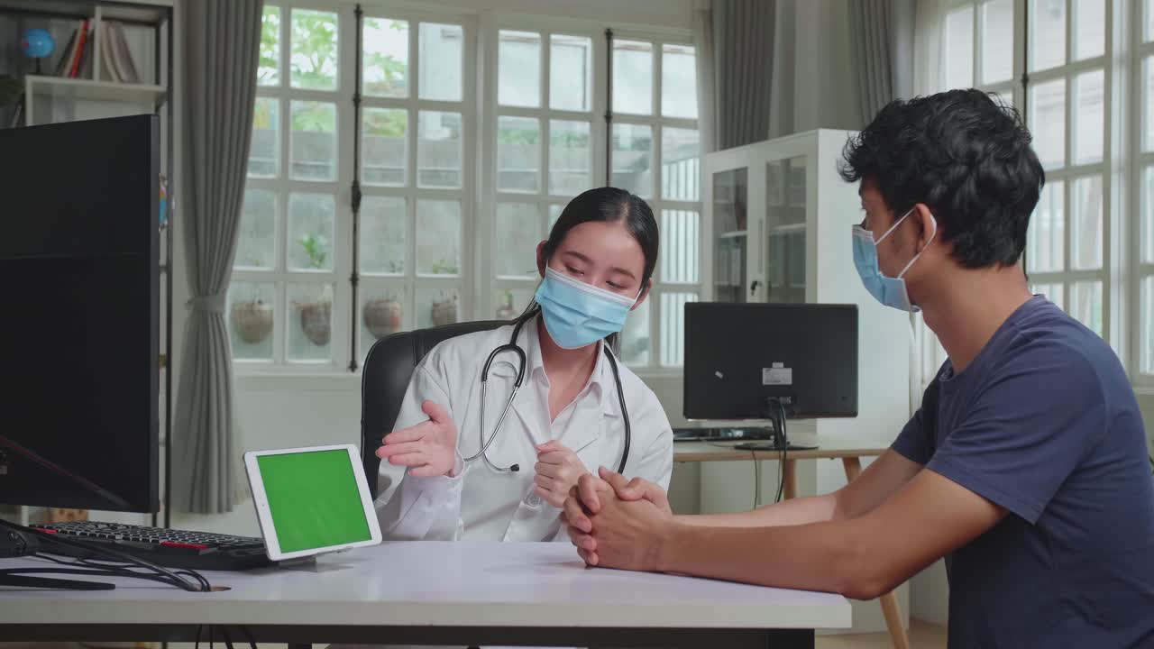 Asian Woman Doctor Showing Green Screen Display On Tablet Computer During Consultation In A Health Clinic. Both Wear Face Masks. Physician In Lab Coat Sitting Behind A Computer Desk In Hospital Office
