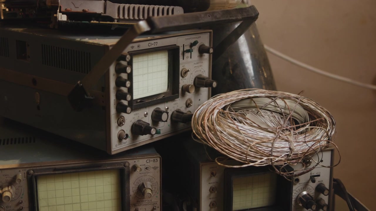 Vista frontale a tre quarti di vecchi strumenti di test elettronici con display a griglia luminosi, manopole, interruttori, cavi, patina, polvere e superfici usurate in un ambiente di officina pratica, con oscilloscopio, CRT