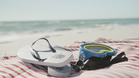 Close-up-of-goggles,-flip-flops-and-towel-on-beach,-in-slow-motion,-with-copy-space