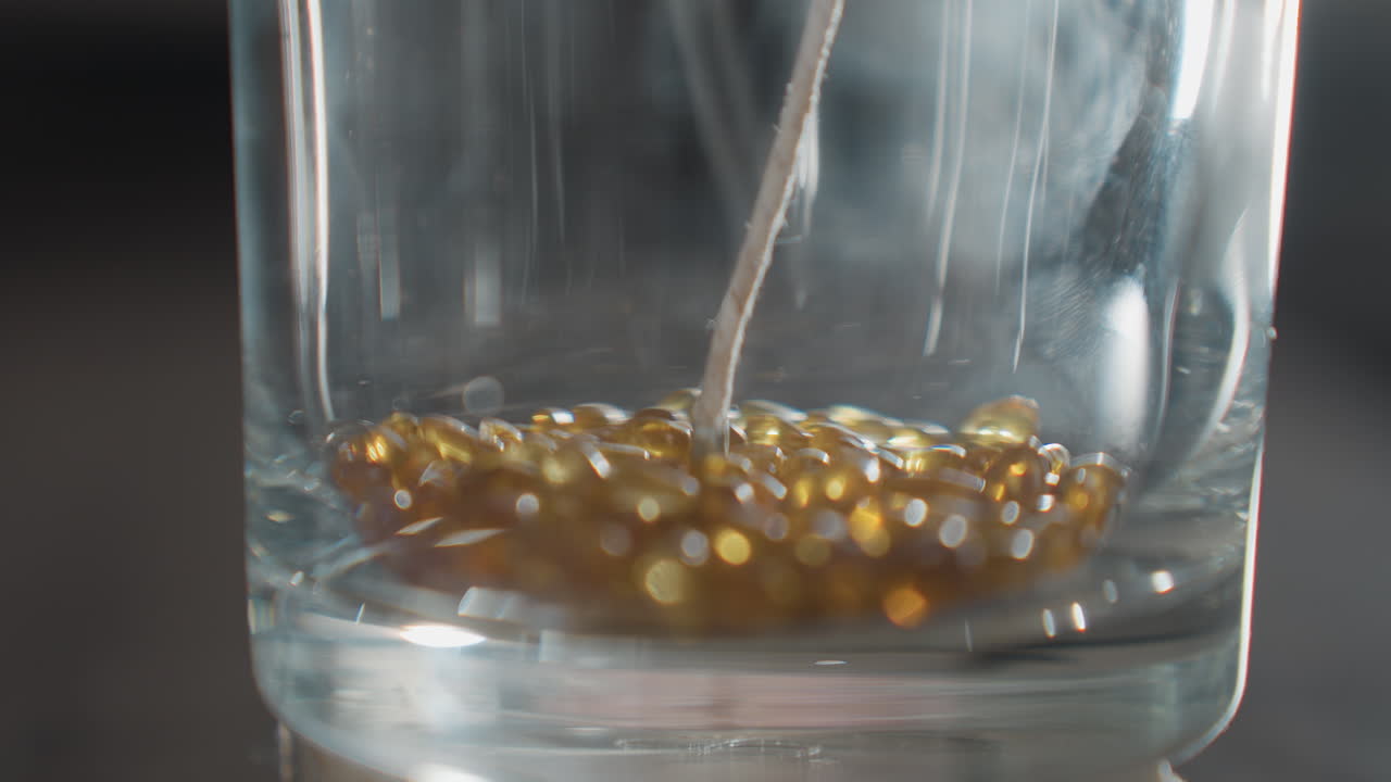 Close up of transparent glass cup with white wick as golden brown beads gently fall inside creating dynamic motion, with beads scattering lightly across glossy surface in soft natural lighting
