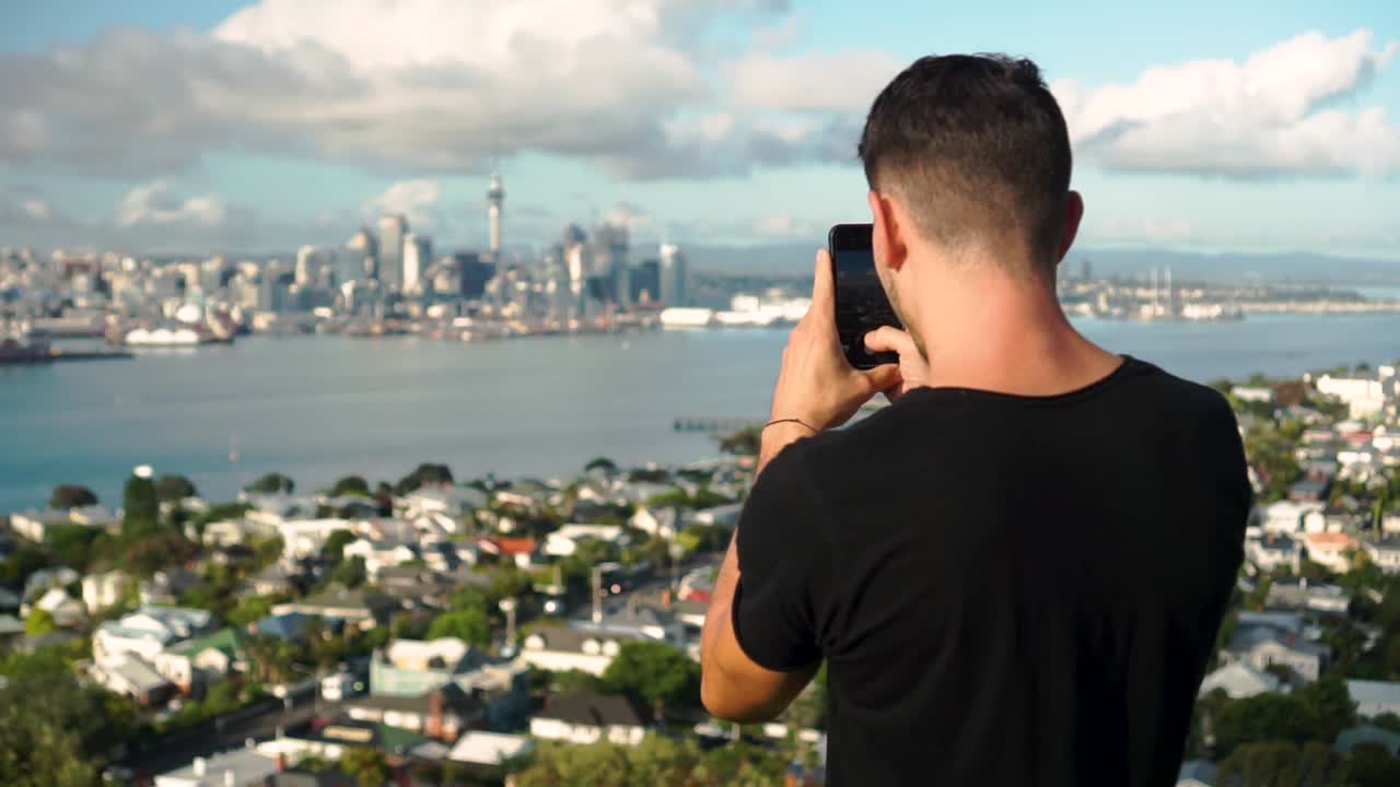 slowmo - joven turista caucásico tomando una foto con su teléfono de auckland y sky tower desde el monte victoria, nueva zelanda