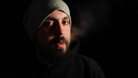 Man-standing-in-dark-room-smoking-cigarette-and-looking-to-camera-very-stressed-and-frustrated