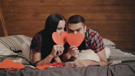 adult man and woman in plaid shirts are lying on the bed and peeking out from under red paper hearts in their hands in the studio. valentine's day. love story