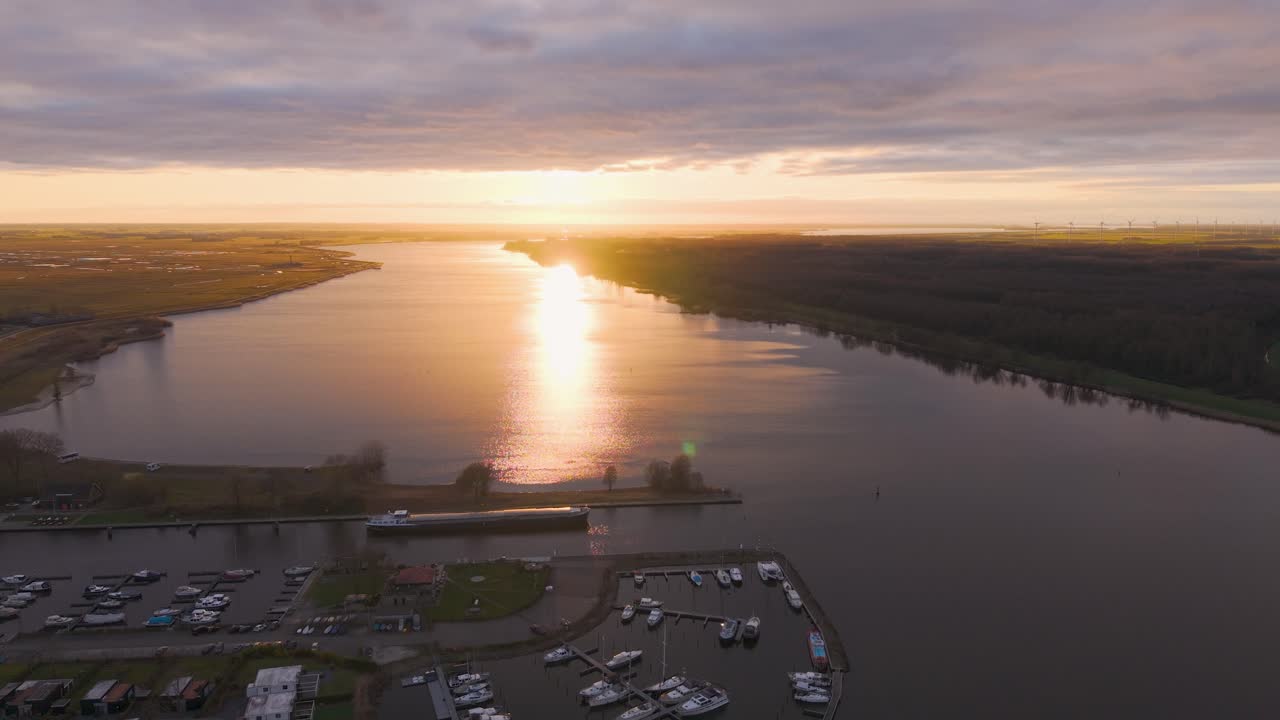 Drone view of a sunset over a river with a marina, boats, and wind turbines in the distance, reflecting golden light.