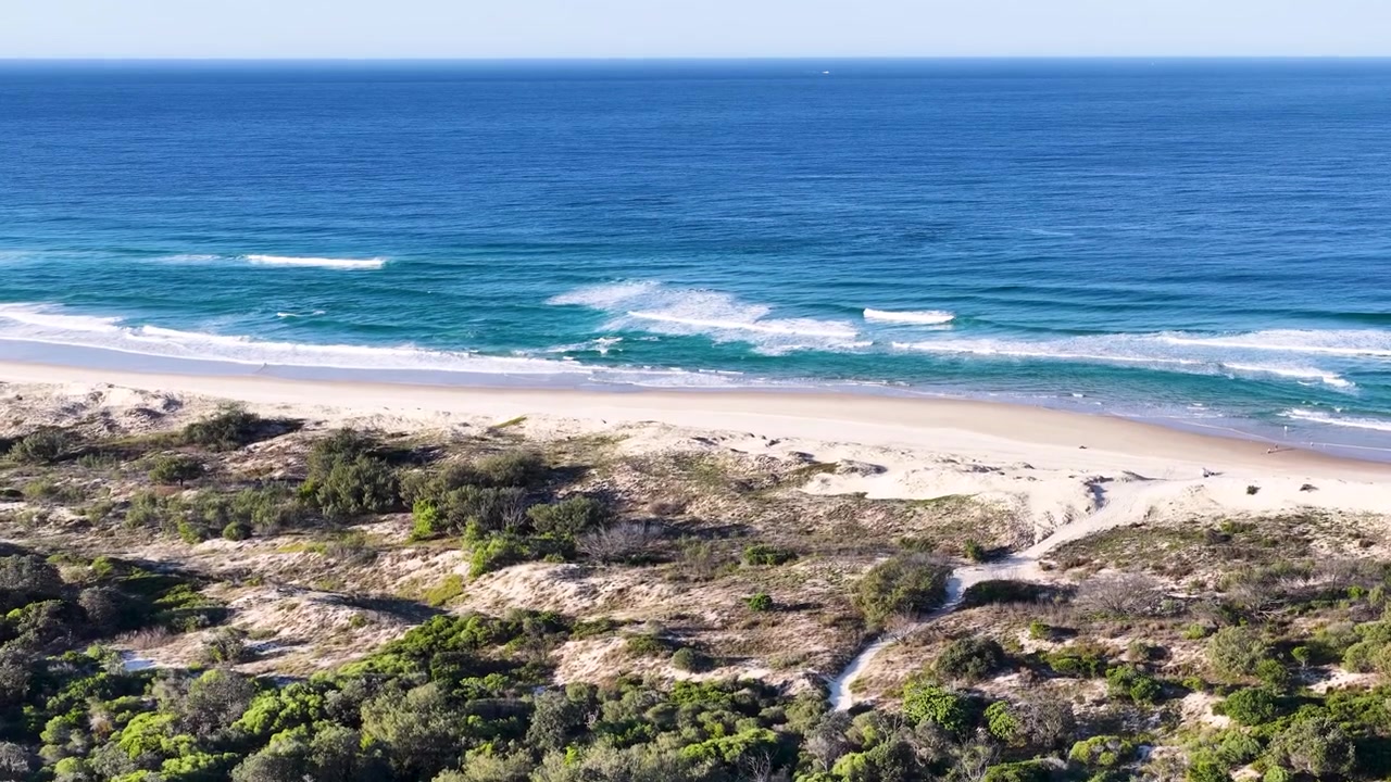 Aerial wide shot of sand dunes, coastal vegetation, and ocean waves at South Stradbroke Island.