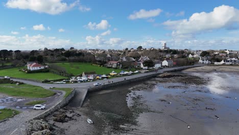 Reveal-of-southside-of-Bordeaux-Harbour-Guernsey-at-mid-tide-with-harbour-wall,-boats-on-hardstanding,beach-and-cottages-in-the-background-on-a-bright-day