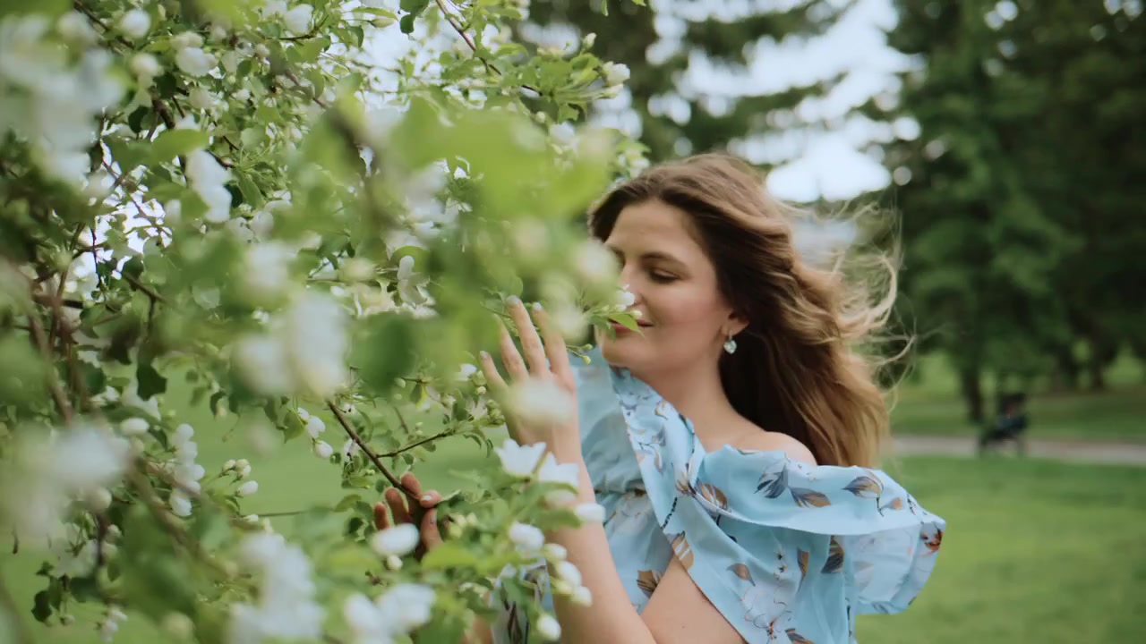 Una donna sorride dolcemente, toccando delicatamente i rami fioriti di un albero. Indossa un vestito floreale azzurro, i capelli le ondeggiano al vento. La scena appare calma e naturale, perfetta per progetti primaverili o estivi.