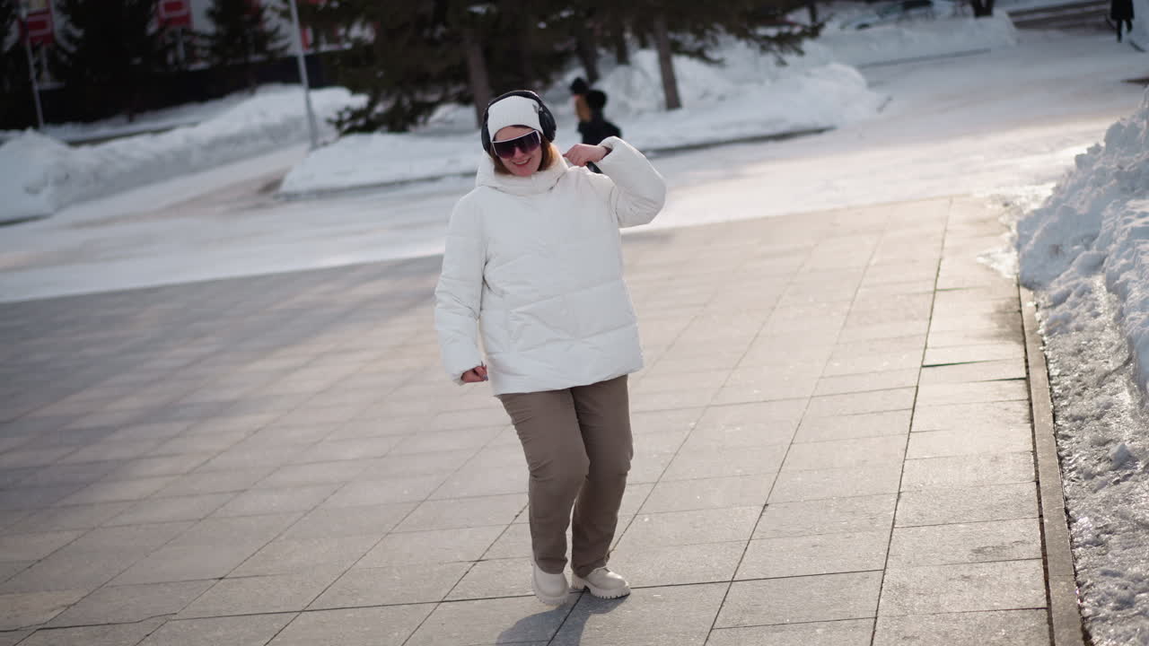 Closeup of dancer wearing white puffer jacket, beanie, goggles and headphones practicing dance moves on large tiled plaza with faint passersby on snowy ground and trees lining background