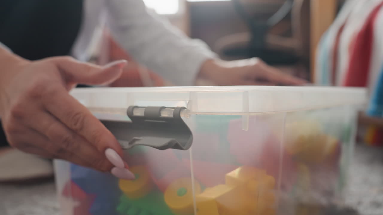Closeup hand closing toy box, mother and son tidy up in sunlit livingroom, colorful building blocks visible through translucent plastic container, gentle manicure on fingers, careful latch click, warm