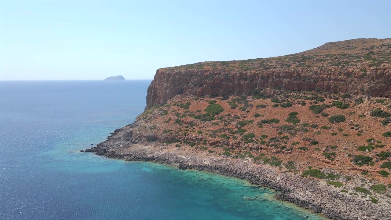 Yunanistan'ın Girit adasındaki Balos burnunun devasa kayalık falezlerini sergileyen nefes kesici drone çekimi. Derin turkuaz ve mavi deniz suyuna, ufukta küçük, uzak bir adaya ve parlak güneşli bir güne sahip.