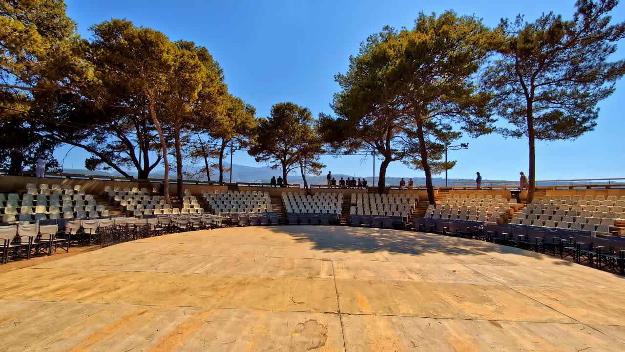 Open-air theater with wooden stage surrounded by trees and empty seats under a bright blue sky