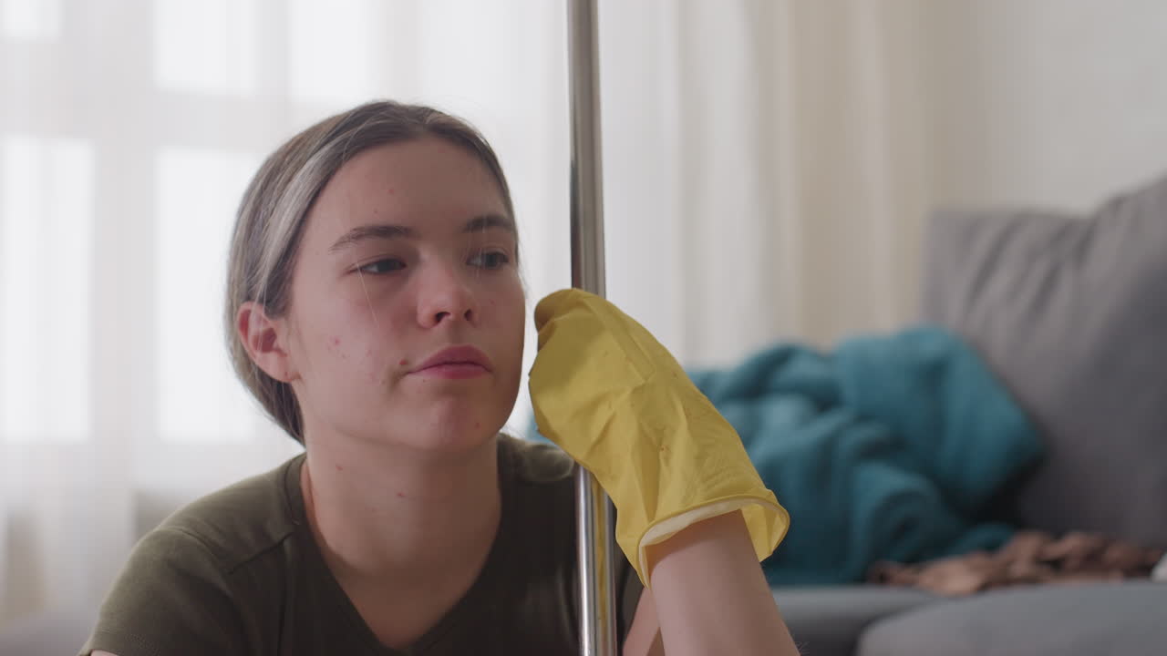 Close up of young woman wearing yellow gloves holding mop handle thoughtfully with tired expression, resting her head against handle while sitting in cluttered living room during household cleaning
