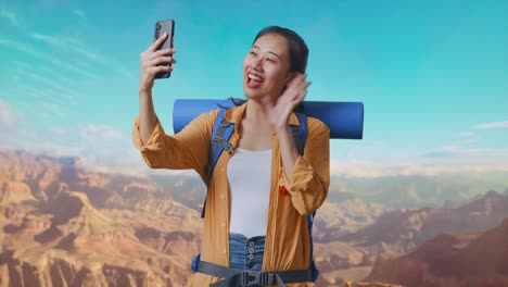 asian female hiker with mountaineering backpack waving her hand while having a video call on smartphone while traveling at the top of mountain