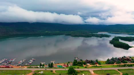 Aerial-Drone-View-of-Shadow-Mountain-Reservoir-and-Grand-Lake-with-Beautiful-Reflection-of-Mountains-and-Rolling-Coulds-as-Cars-Drive-on-Colorado-Highway-Road-Along-the-Overcast-Shoreline