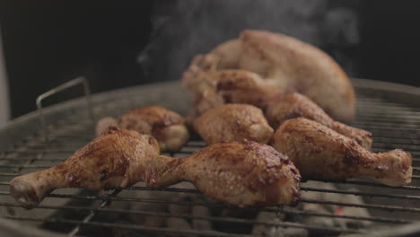 chicken drum sticks getting grilled on coal and fire with smoke coming up with black background and a whole chicken in the back shot raw and 4k eye level