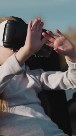 niña preescolar con discapacidad mira las manos a través de gafas vr. mujer en silla de ruedas entretiene cerca del estanque en un día soleado en un primer plano de fondo borroso