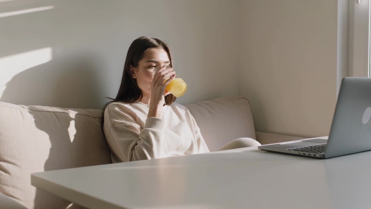 Kigger på bærbar computer, siddende kvinde i let sweater nipper til appelsinjuice på sofaen under arbejdsfrokost
