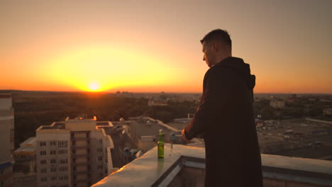 close the lid of the laptop and drink beer standing on the roof. a man in a hoodie with a laptop in his hands at sunset.