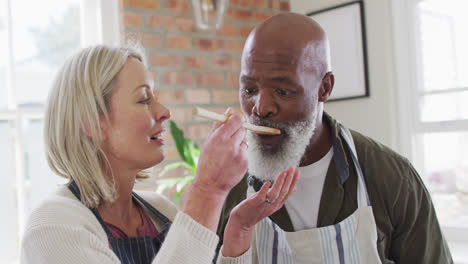 Pareja-De-Ancianos-De-Raza-Mixta-Usando-Delantales-Probando-Comida-Mientras-Cocina-En-La-Cocina-De-Casa