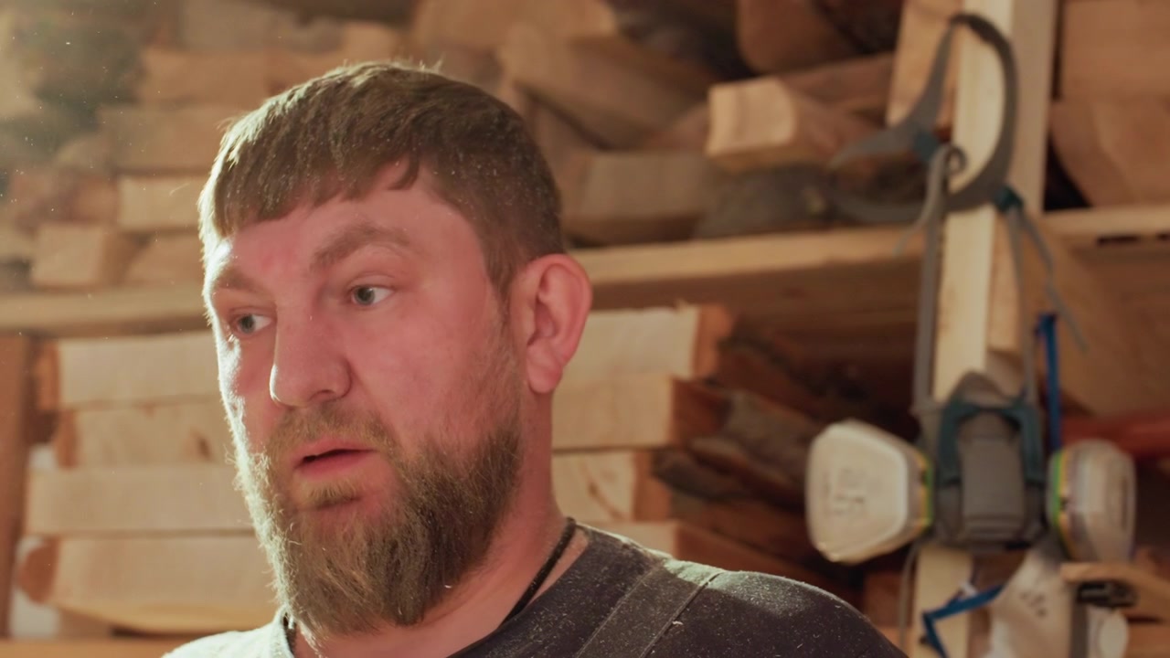 Close up shows bearded woodworker covered in sawdust speaking directly to camera. background reveals stacks of raw timber woodworking equipment and tools within workshop setting