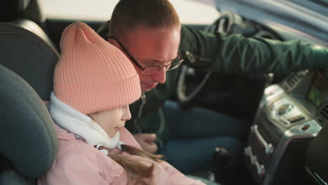 close-up of a young girl smiling slightly, in a pink beanie and jacket, sitting quietly in a car, with a man, likely her father, leaning in closely and attentively looking at the phone she is holding