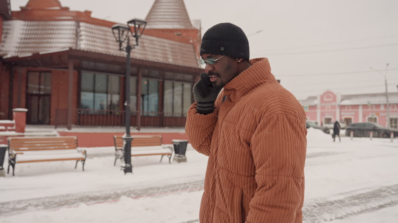 Black man in orange jacket speaking on phone with urgent tone, carrying black backpack, gesturing with gloved hand, brisk steps across snowy sidewalk, urban station backdrop, startup founder closing