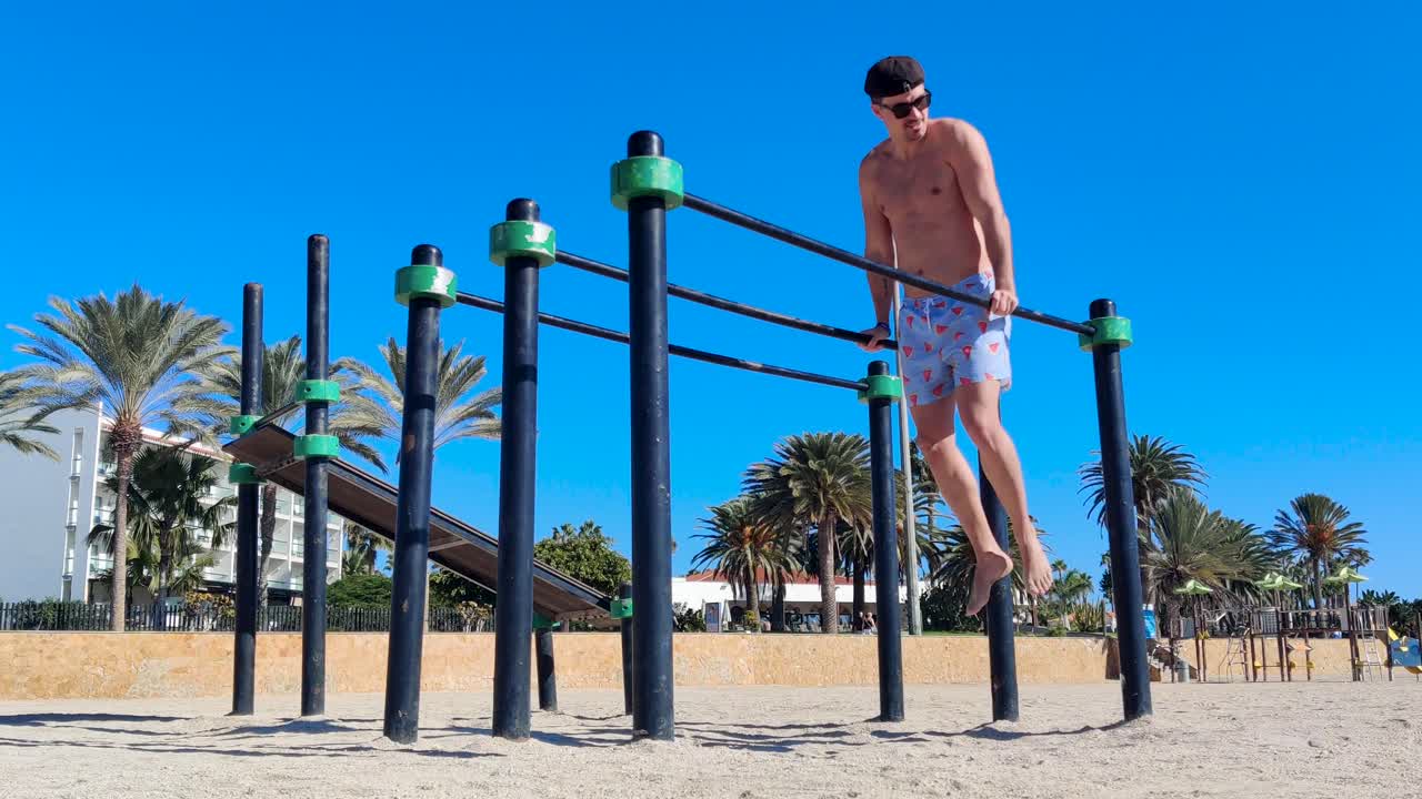 un hombre joven con pantalones cortos, un sombrero de béisbol y gafas de sol haciendo saltos de tríceps en barras paralelas en un gimnasio al aire libre en la playa