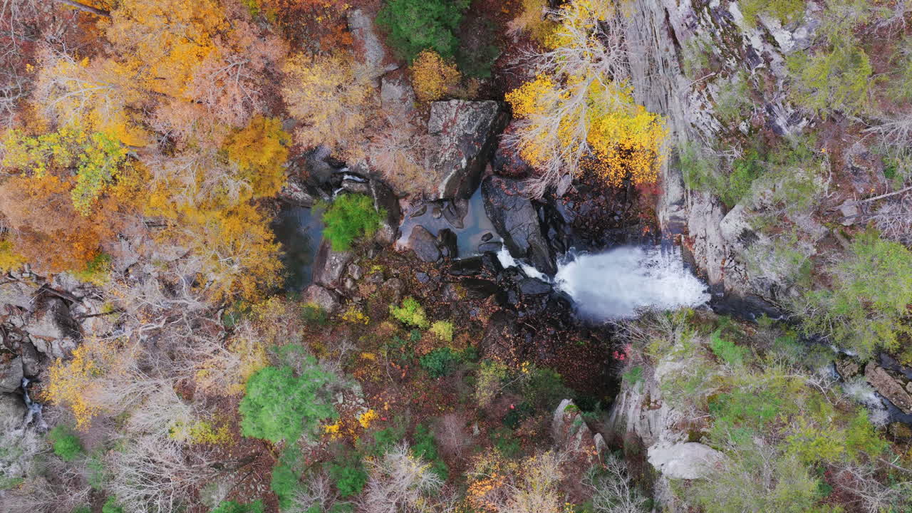Korkea drone-näkymä, jossa näkyy putouksen alaosan allas ja ympäröivä kivinen maasto yhdistettynä hillittyyn syksyn lehdistöön