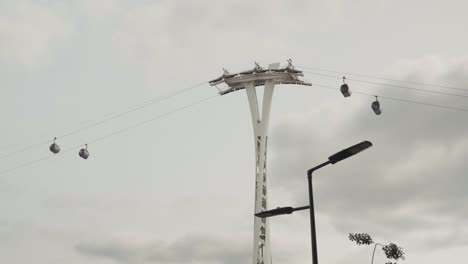 A-modern-urban-cable-car-system-in-London,-featuring-sleek-gondolas-suspended-from-a-tall,-white-support-tower-against-a-cloudy-sky