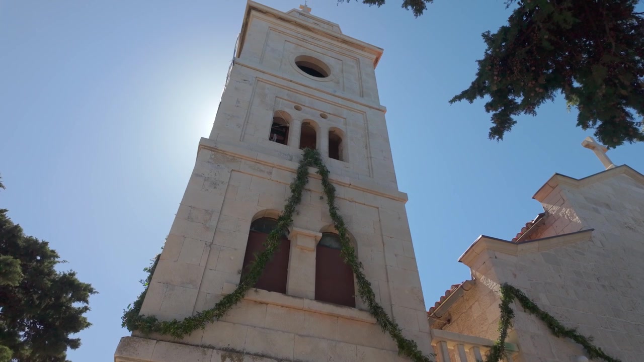 Ripresa aerea ascendente del campanile di San Giorgio a Primošten con il sole che splende dietro, mostrando la chiesa decorata con una ghirlanda verde sotto una luminosa luce costiera.