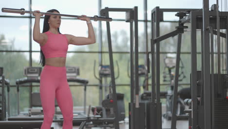 mujer morena hispana con un traje rosa empuja una barra sobre su cabeza hacia arriba mientras entrena sus hombros en el gimnasio. ejercicio de pie para entrenar los hombros y los brazos. entrenamiento con pesas