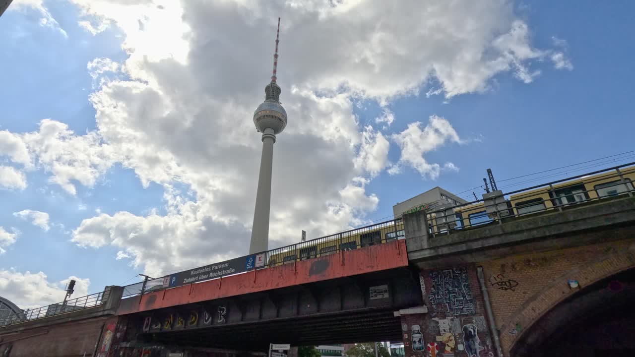 A yellow and red commuter train crosses a graffiti-covered bridge beneath the Berlin TV Tower, with dynamic clouds and bright daylight. Slight upward camera angle