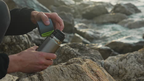 man-unscrews-thermos-and-pour-hot-coffee-into-cup-as-sea-waves-wash-over-rocks-in-background-during-golden-hour