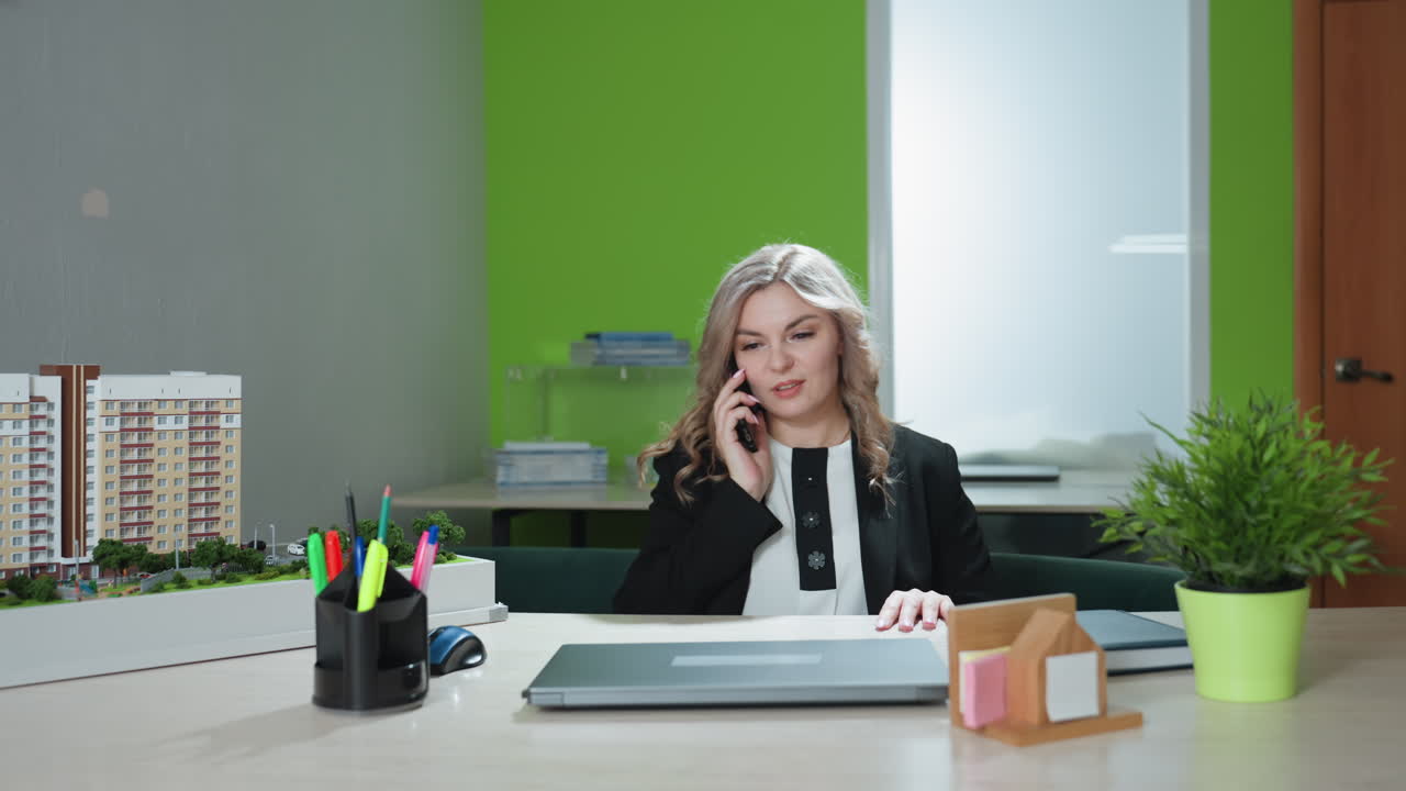 Realtor engaged in client phone call stands in modern office preparing to multitask while planning her day, surrounded by desk with documents and supplies, poised to begin work on laptop